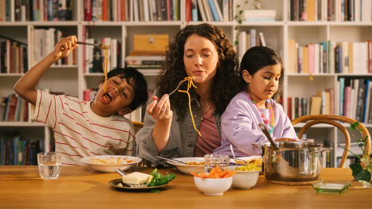 Bild på mörkhårig kvinna med två barn, en flicka och en pojke. De sitter vid ett bord och äter pasta.