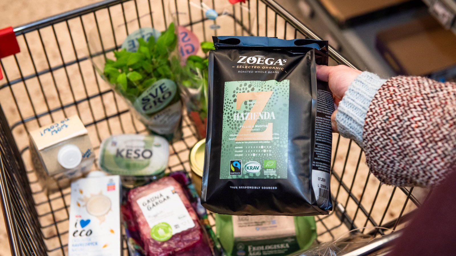 En hand lägger när en påse med Zoegas KRAV-märkta kaffe i en kundvagn där det redan ligger en del varor.